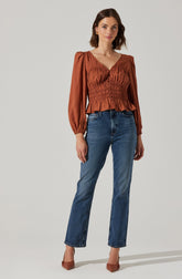 A woman is wearing a rust-colored blouse with long, puffed sleeves and a fitted, ruched bodice with a ruffled hem. She has paired the blouse with high-waisted, straight-leg blue jeans and pointed-toe heels in a matching rust color.